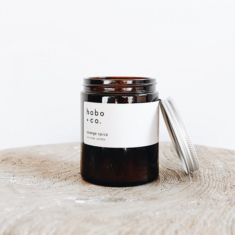 Braunes Glas mit weißem Etikett und silbernem Deckel, enthält eine Duftkerze von hobo + co. auf einem Holzuntergrund.