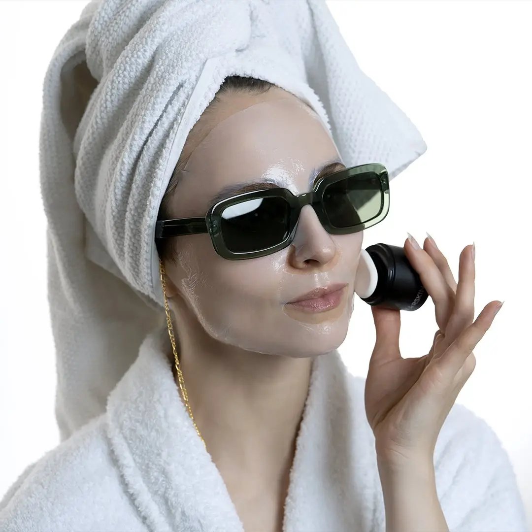 Frau mit Handtuch auf dem Kopf und Sonnenbrille trägt Gesichtsmaske auf.