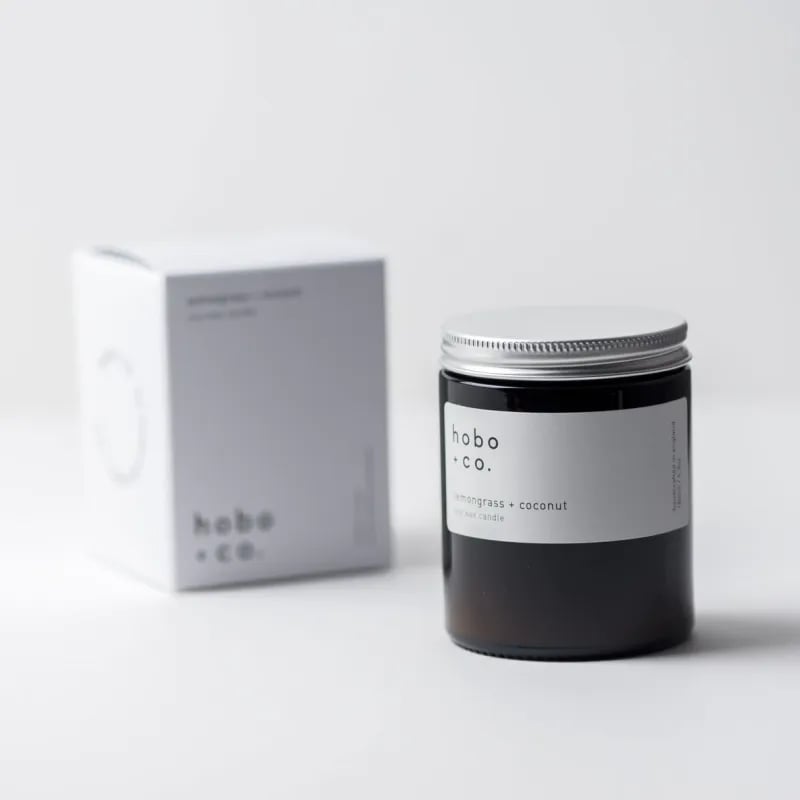 Eine Duftkerze von hobo + co. mit Zitronengras und Kokosnuss, neben der Verpackung auf einem weißen Hintergrund.
