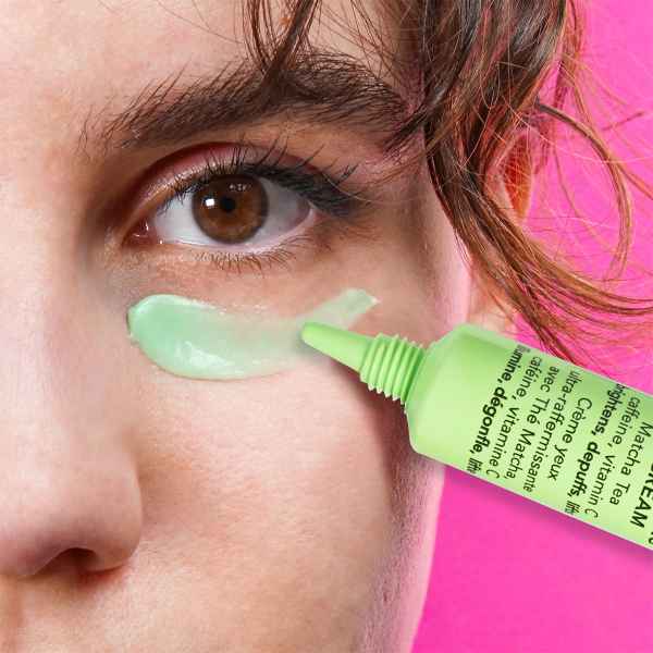 Nahaufnahme eines Auges mit aufgetragener grüner Creme aus einer Tube auf rosa Hintergrund.